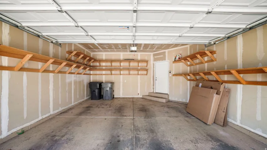 Spacious garage with concrete flooring, overhead garage door, built-in wooden shelving, and a white interior door with steps