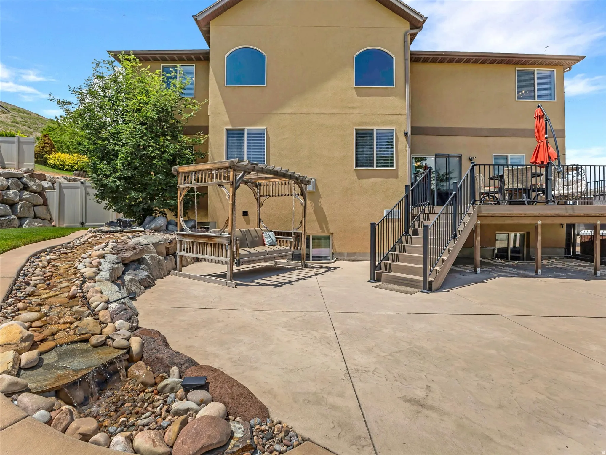 Expansive concrete patio featuring a rock-lined water feature, a wooden pergola, and a multi-level deck with black railings
