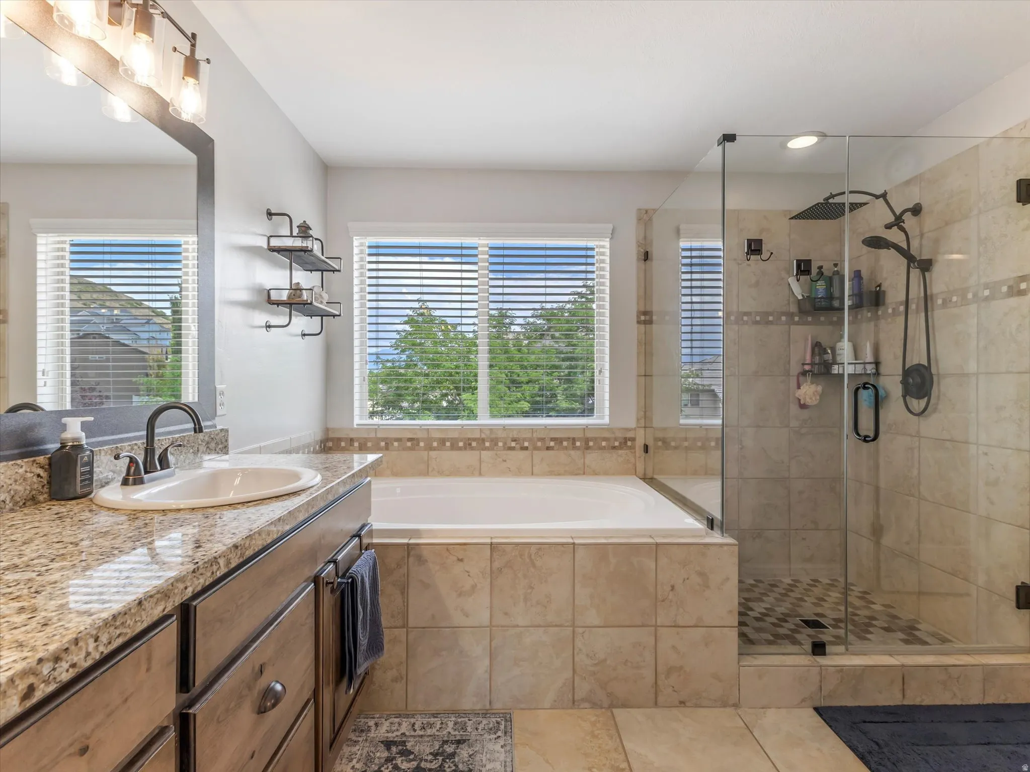 Bathroom featuring a built-in soaking tub, a glass-enclosed shower with a rainfall showerhead, a single vanity with a granite-look countertop and under-sink cabinetry, and two windows with blinds