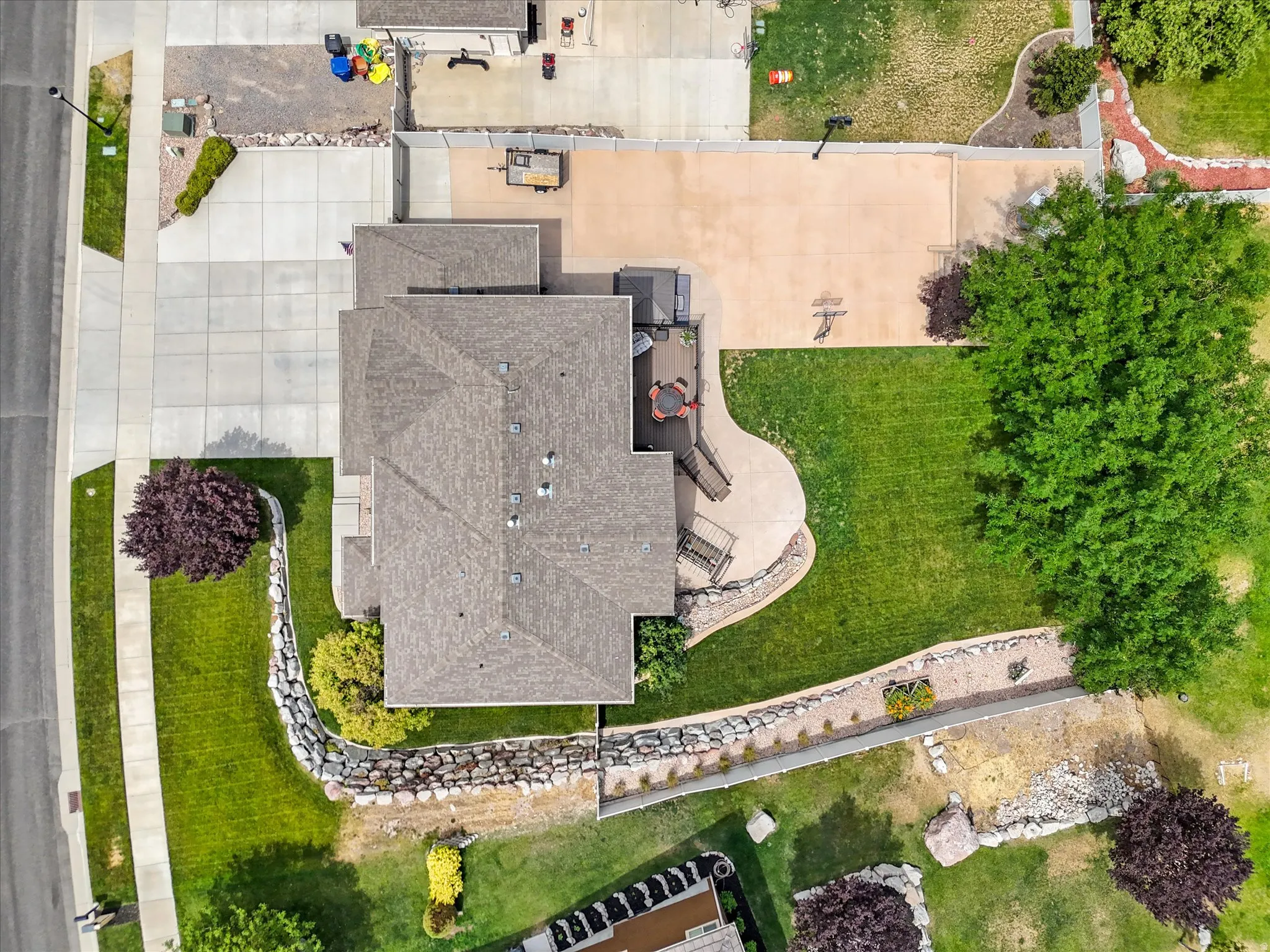 Property featuring a multi-level roofline, extensive paved driveway, a large rear deck with outdoor seating, a curved patio, and a spacious lawn bordered by retaining walls