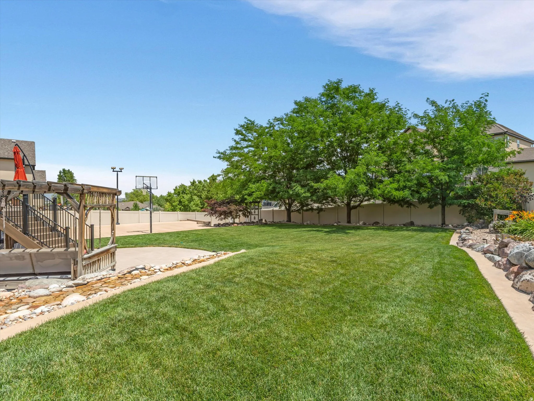 Expansive backyard featuring a manicured lawn, a wooden deck with a pergola, a hardscape patio, a basketball court, and mature trees