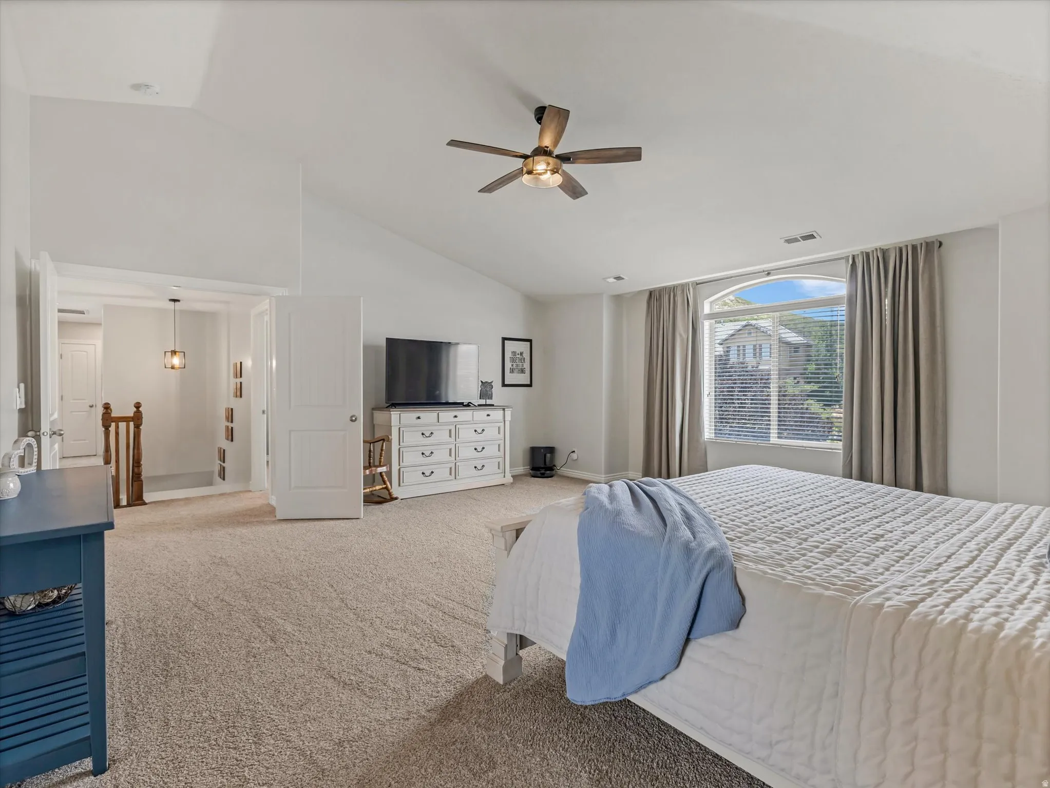 Spacious bedroom featuring vaulted ceilings, neutral-toned carpet flooring, a ceiling fan, and a large arched window with blinds and drapes