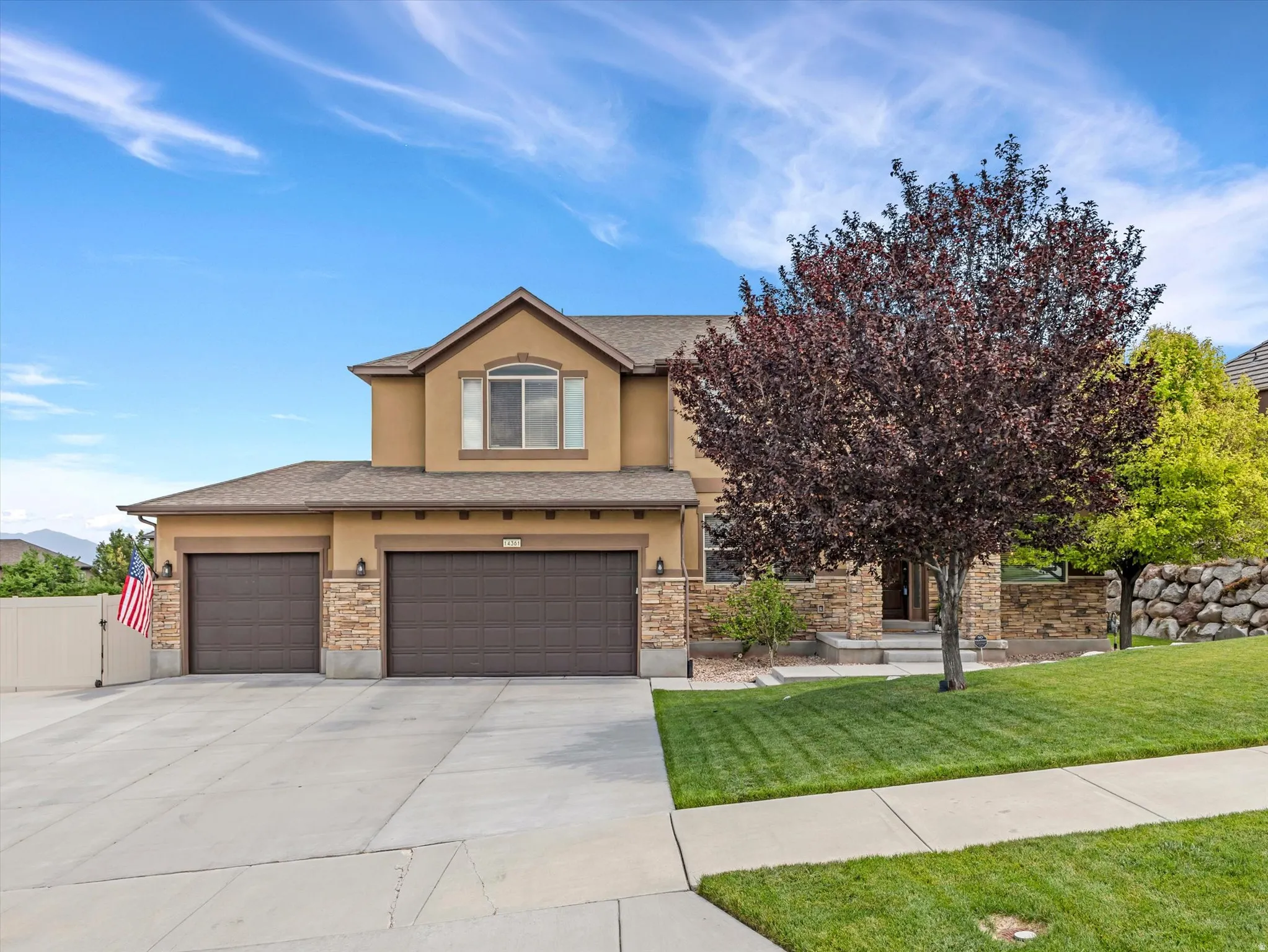 Two-story stucco and stone-accented residence featuring a two-car garage, mature front yard tree, and an arched window on the upper level