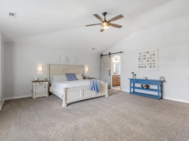 Spacious room featuring vaulted ceilings, a ceiling fan with light fixture, wall-mounted sconce lighting, and a barn door entry to an additional space