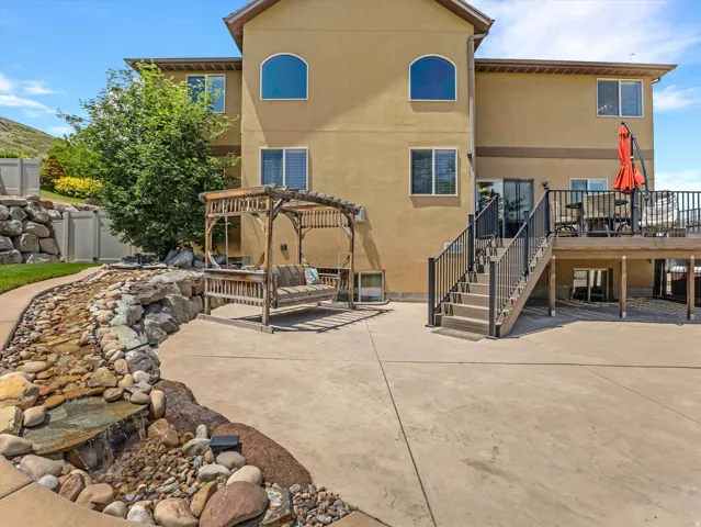 Expansive concrete patio featuring a rock-lined water feature, a wooden pergola, and a multi-level deck with black railings