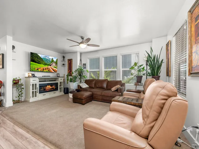 Open-concept living space featuring neutral carpet flooring, a built-in entertainment console with an electric fireplace, multiple windows with horizontal blinds, and a ceiling fan with integrated lighting