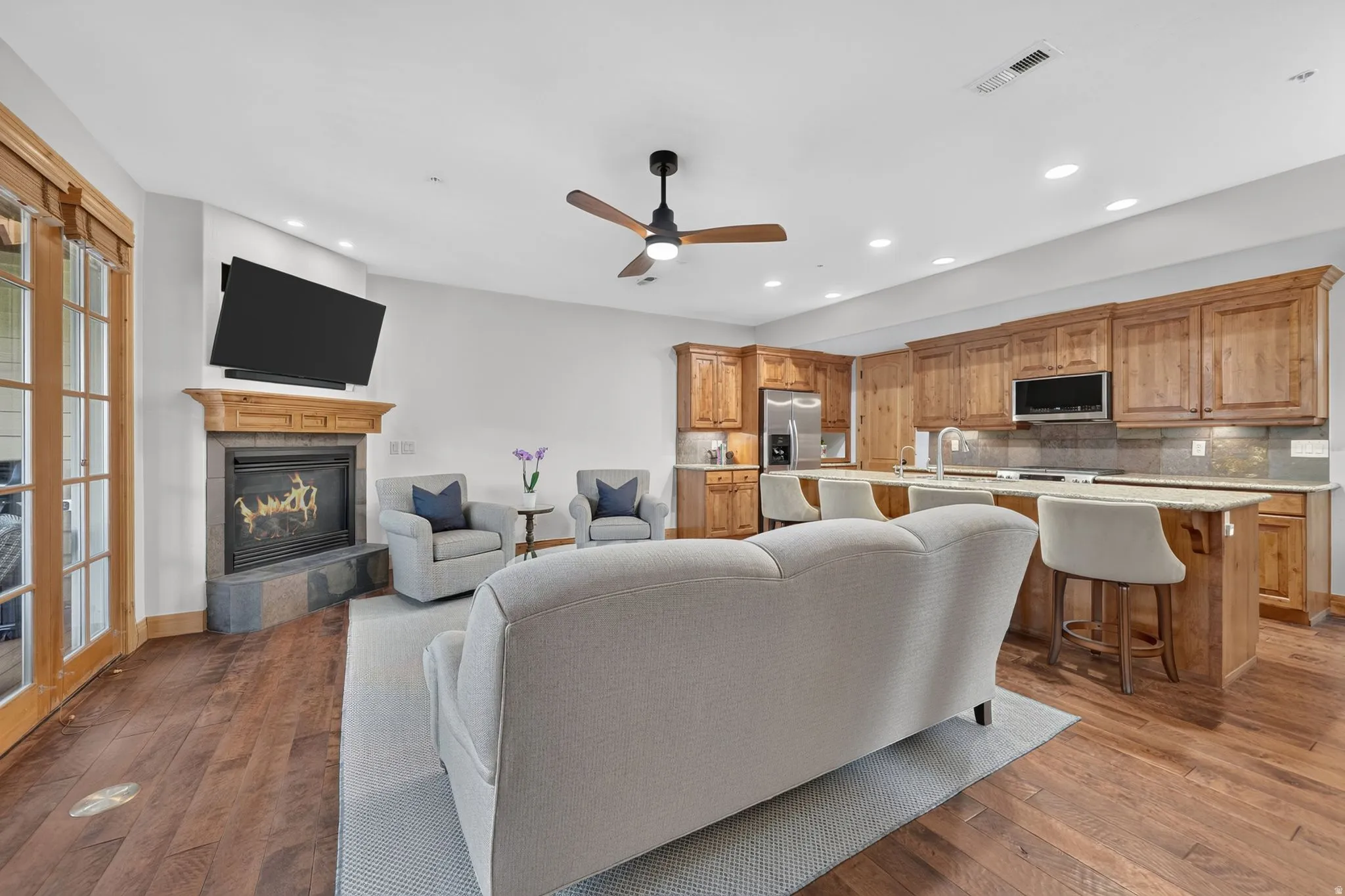 Open-concept living space featuring wood-finish flooring, a stone-surround fireplace with a wood mantel, a ceiling fan, and recessed lighting