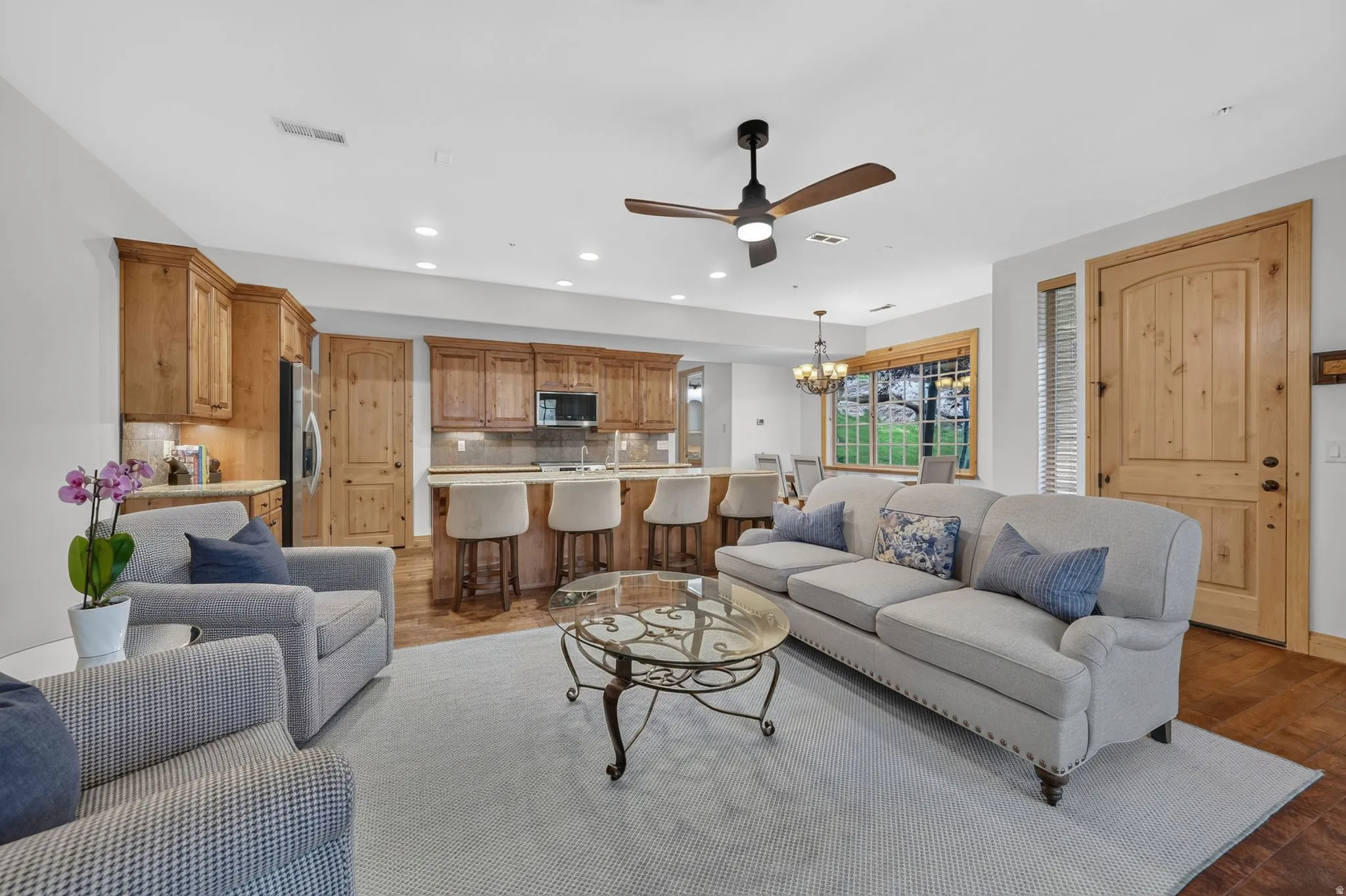 Open-concept living space featuring wood-finish flooring, a ceiling fan with light fixture, and a kitchen with wood cabinetry, a built-in microwave, and an island with counter seating