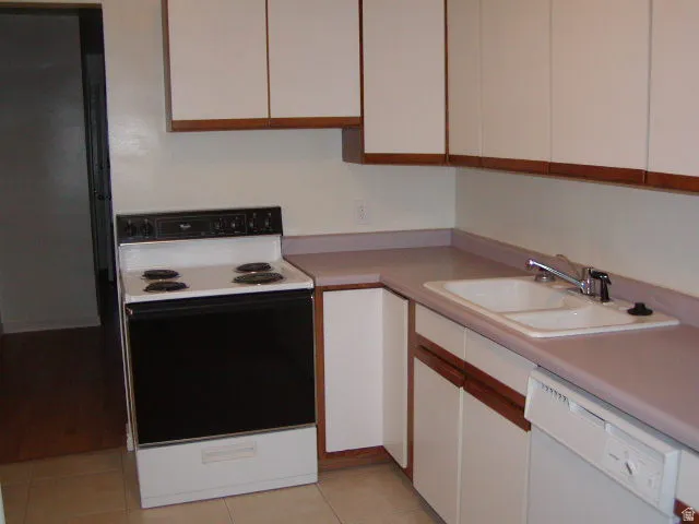 Kitchen featuring white cabinetry with wood-finish trim, a white electric range, a double basin sink with chrome faucet, a white built-in dishwasher, and a light-toned countertop