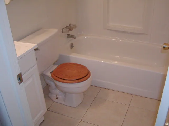Bathroom featuring a white bathtub with integrated shower, a toilet with a wood-finish seat, a white vanity with a light-colored countertop, and square tile flooring
