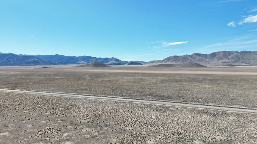 Expansive desert landscape featuring a clear sky, distant mountain ranges, and a visible road