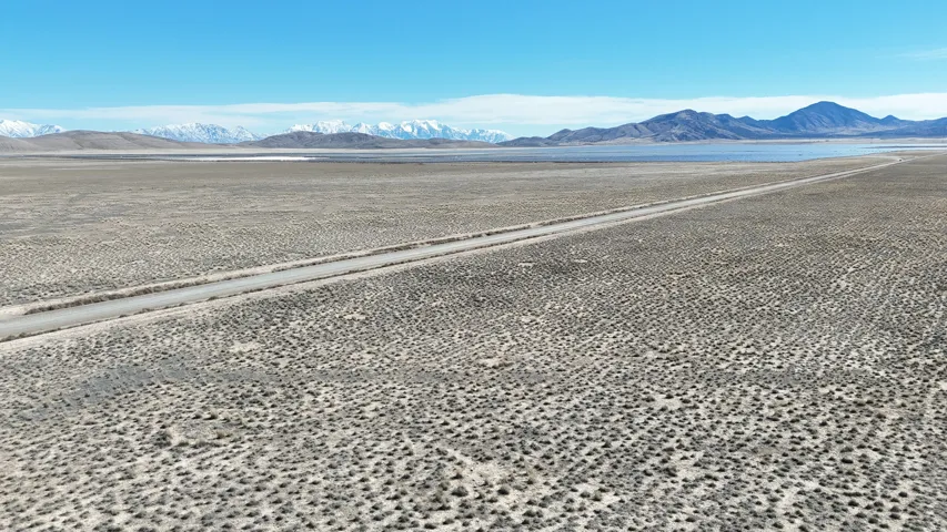 Expansive open landscape featuring a dirt road, distant mountains with snow-capped peaks, and a visible body of water