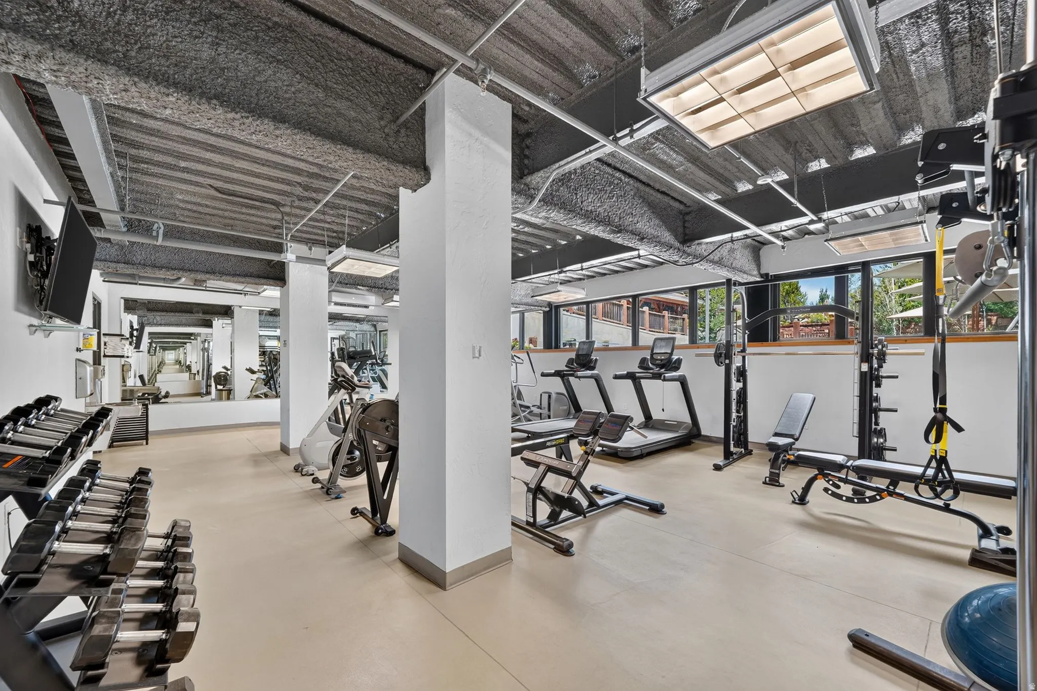 Fitness center featuring multiple exercise machines, free weights, wall-mounted television, full-length mirrors, and ample natural light from exterior windows