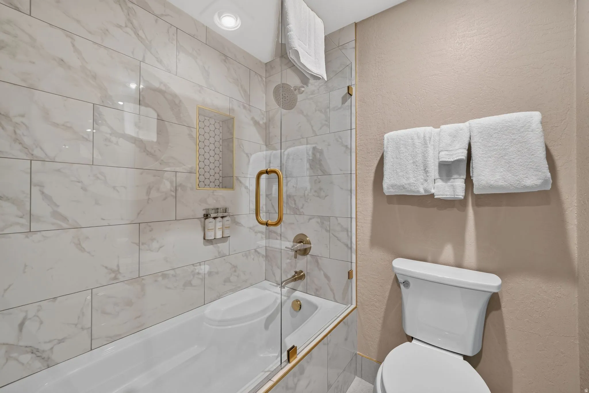 Bathroom featuring a tub and shower combination with a clear glass enclosure, white subway tile with a marble-effect pattern, gold-tone fixtures, and a recessed shower niche with hexagonal mosaic tile