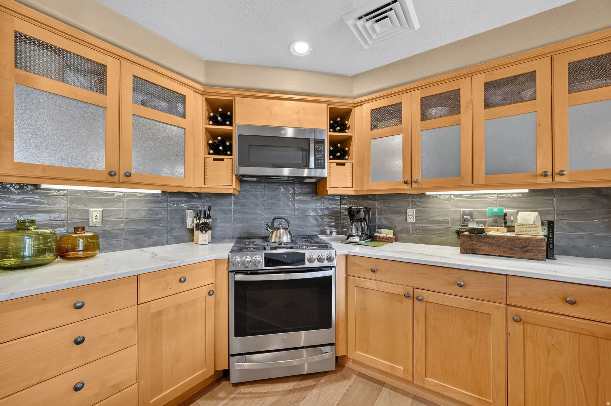 Kitchen featuring light wood cabinetry with frosted glass and metal mesh inserts, stainless steel appliances, a white countertop, and a dark tiled backsplash