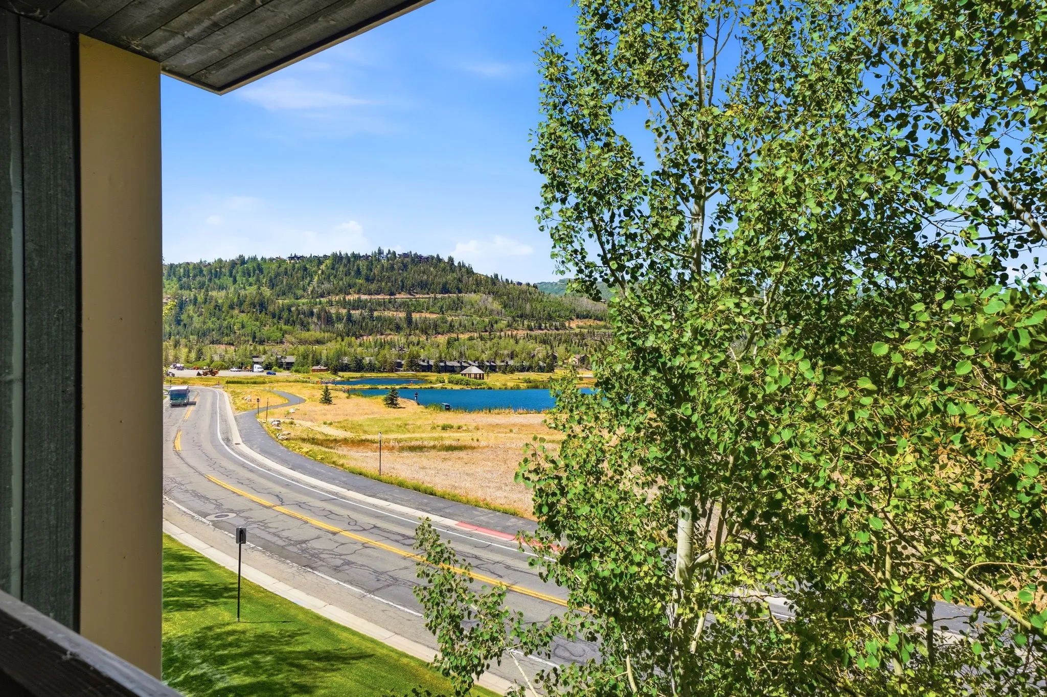 Exterior balcony overlooking a paved road, a serene blue lake, and distant tree-covered hills under a clear sky