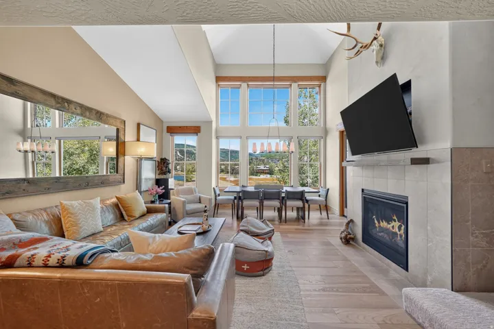 Open-concept living space with vaulted ceilings, large windows, wood-finish flooring, a built-in fireplace with a tiled surround, and a dining area