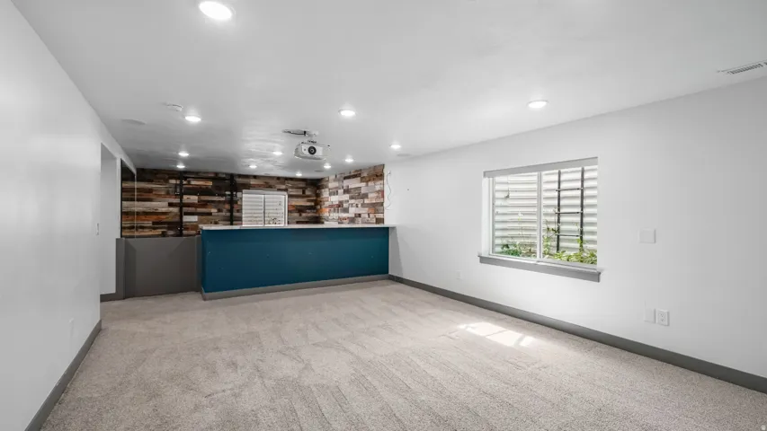 Carpeted recreation room featuring recessed lighting, a built-in bar with a vibrant teal base and wood-finish accent wall, and a window with a window well