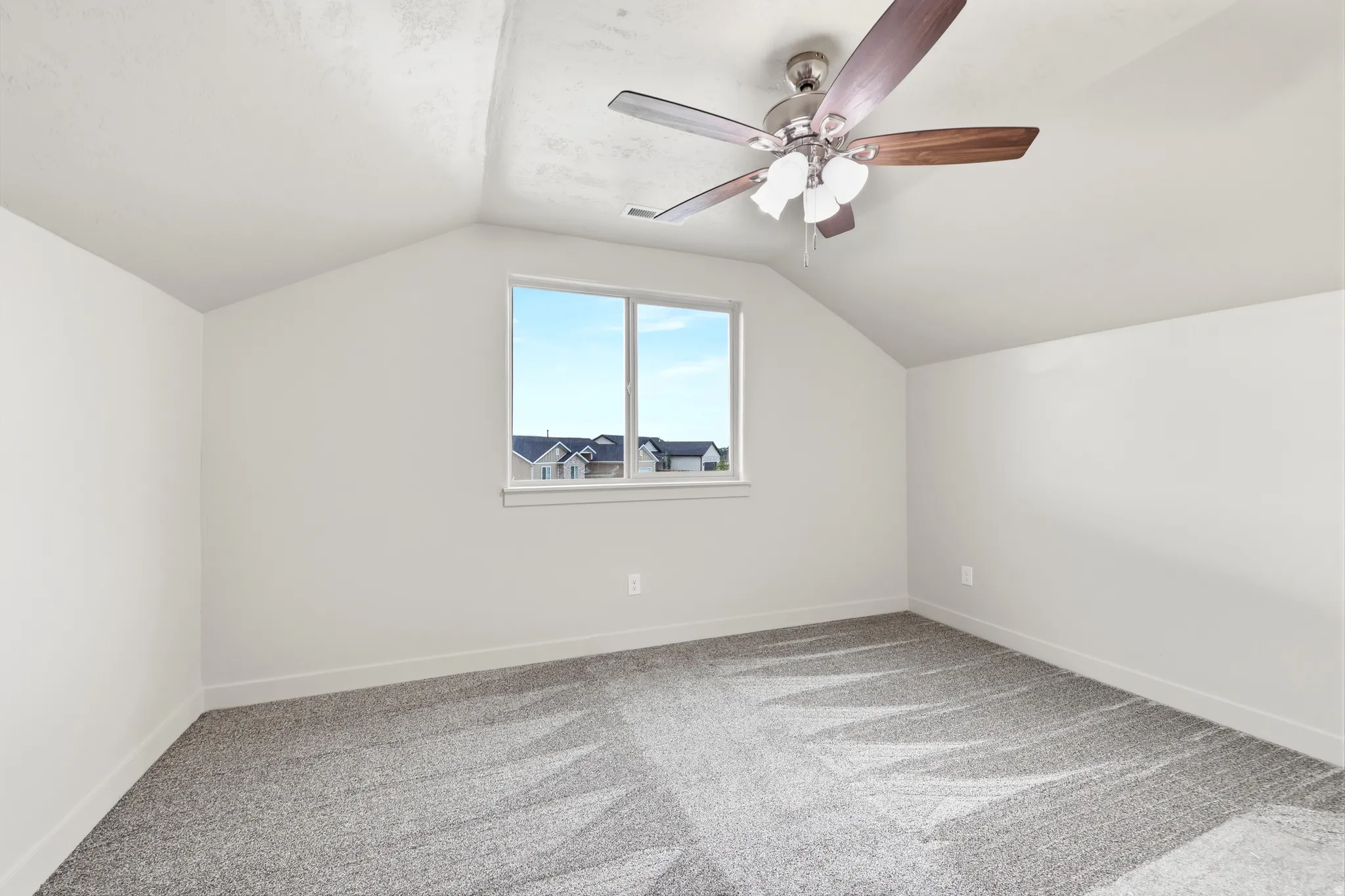 Carpeted room featuring a window with exterior residential structures visible, sloped ceilings, a ceiling fan with light fixture, and white baseboards