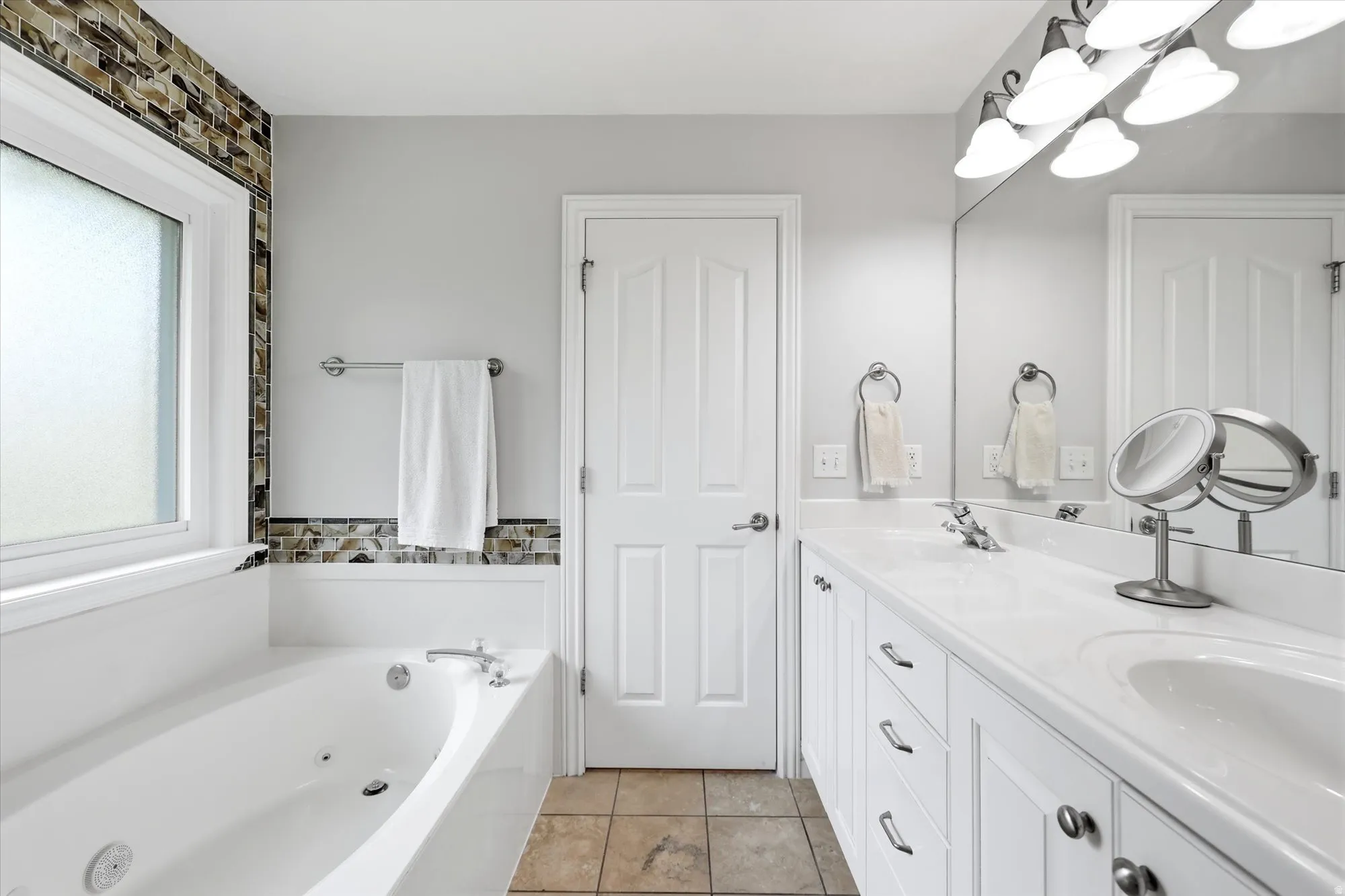 Attached to the primary bedroom. Spacious bathroom featuring a white soaking tub with jets, a mosaic tile accent wall, a double vanity with white cabinetry and integrated sinks, and neutral floor tiling