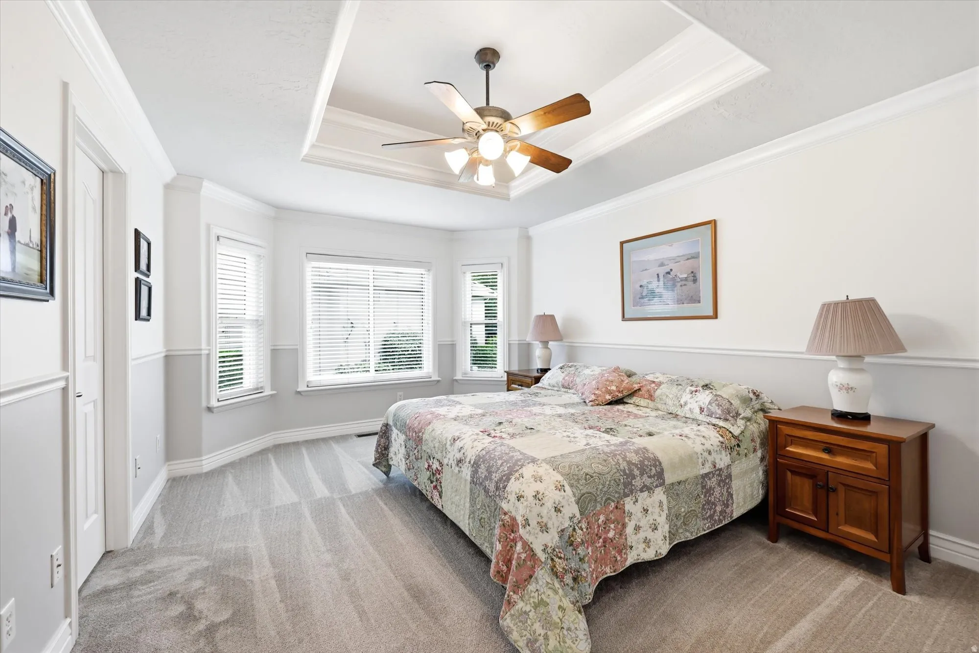 Primary Bedroom. Recessed tray ceiling with ceiling fan, bay window with blinds, soft gray carpeting, white wainscoting, and white trim