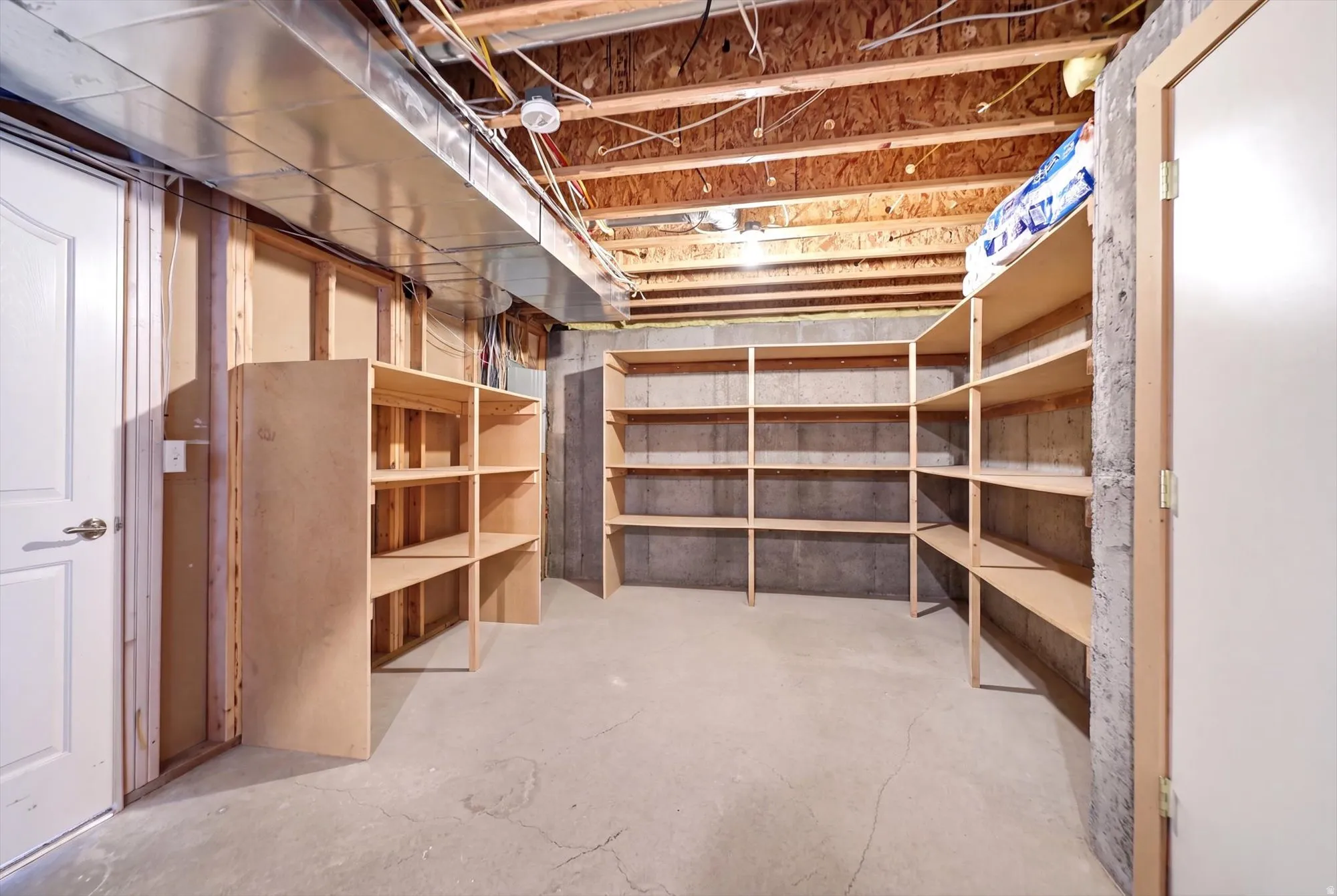 Basement storage area featuring extensive built-in shelving, a concrete floor, and exposed ceiling joists with visible ductwork