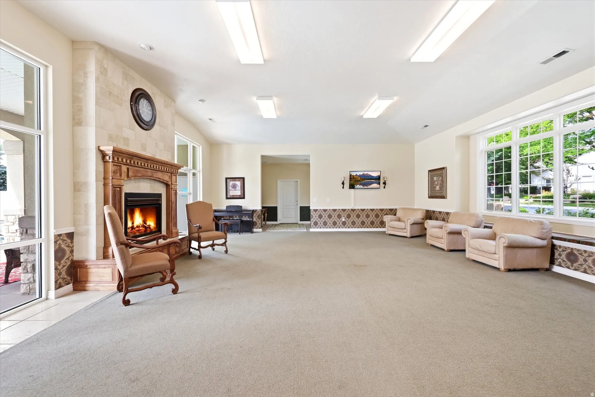 HOA Clubhouse Spacious common area featuring a stone-clad fireplace with a decorative wood mantel, vaulted ceilings, large picture windows, and patterned wainscoting
