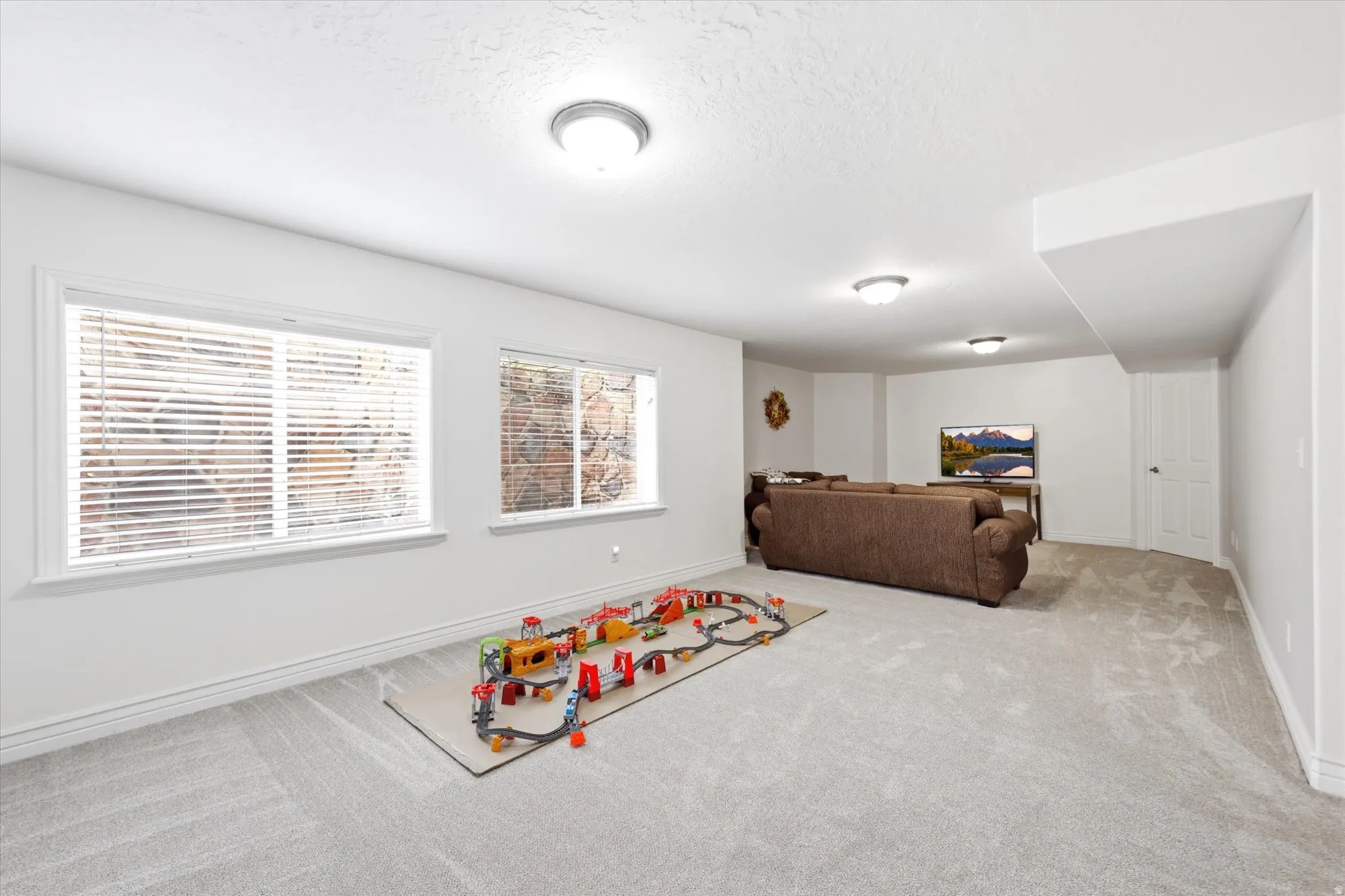 Spacious lower-level room featuring light-toned carpeting, white baseboards, and two windows with horizontal blinds