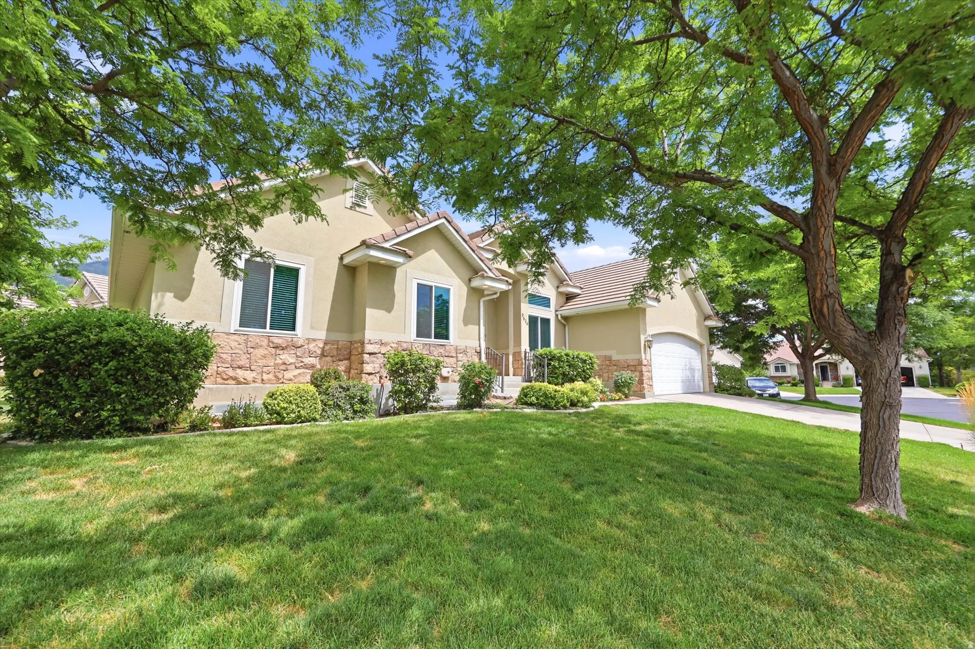 Stucco exterior with a stone veneer accent, mature trees, manicured lawn, and an attached two-car garage