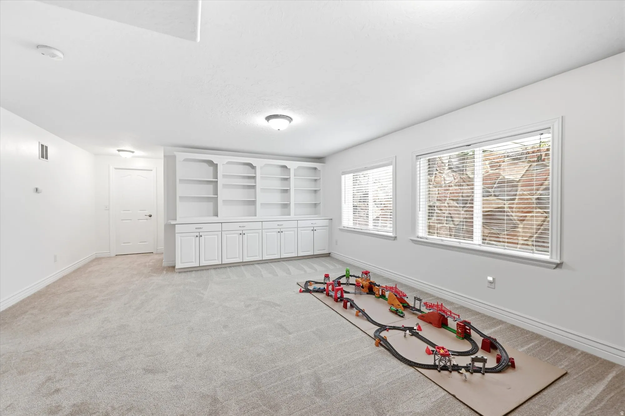Spacious room featuring light-colored carpeting, multiple windows with blinds, recessed lighting, and a built-in shelving unit with lower cabinetry