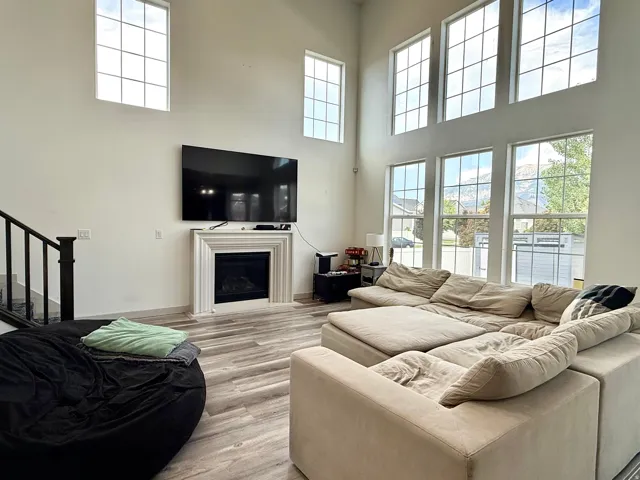 Two-story living area featuring a decorative fireplace with mantel, staircase with dark railing, wood-finish flooring, and multiple tall windows providing abundant natural light