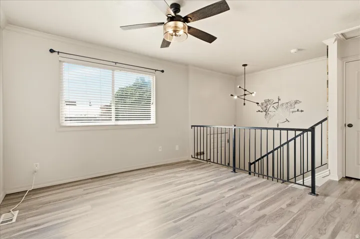 Light wood-finish flooring, ceiling fan with integrated lighting, window with horizontal blinds, black metal railing, and a contemporary chandelier