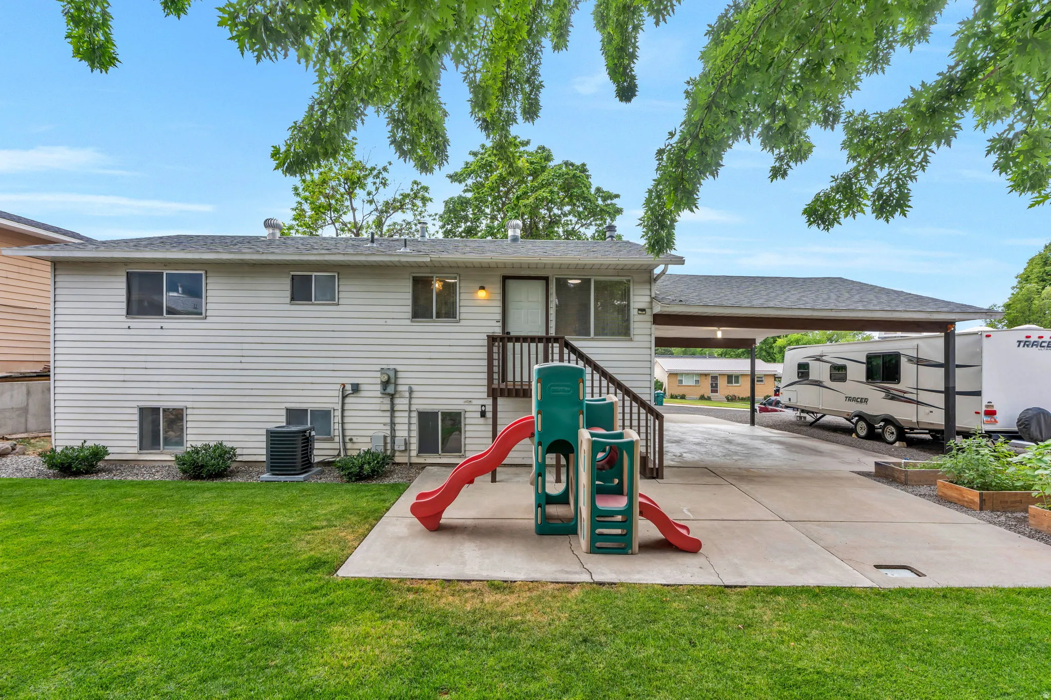 Rear exterior featuring white horizontal siding, a ground-level concrete patio, a multi-level deck with wood railing, a covered carport extension, and multiple raised garden beds