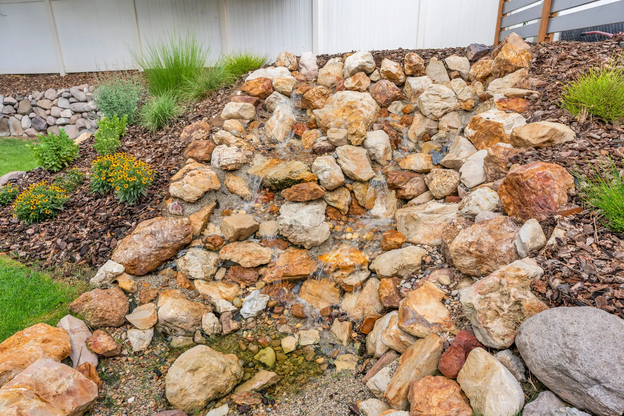 Rock water feature with flowing water, surrounded by natural stone landscaping, mulched garden beds, and manicured green lawn