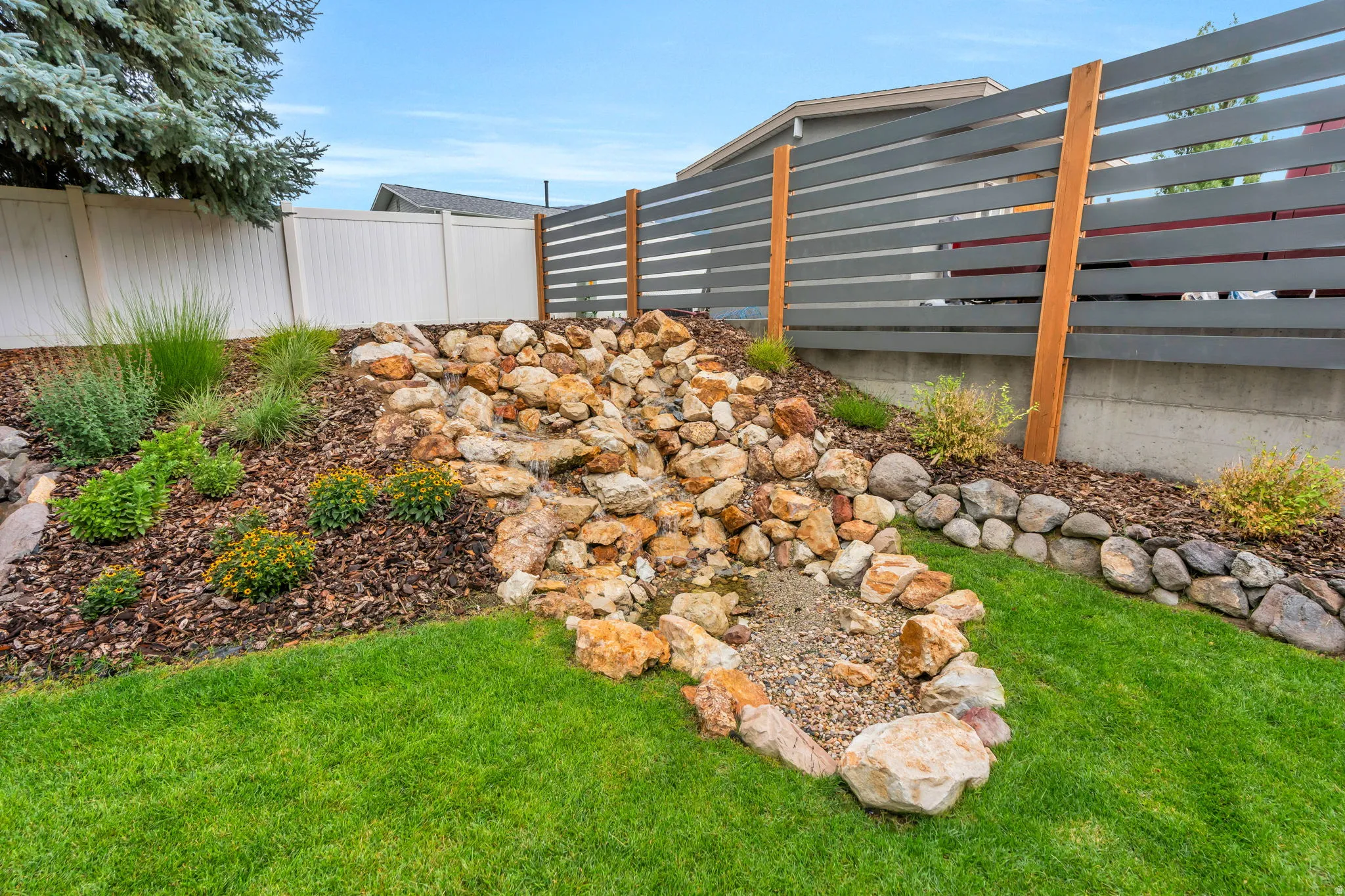 Backyard rock garden featuring a cascading water feature, landscaped planter beds with mulch, a manicured lawn, and a contemporary horizontal slat fence
