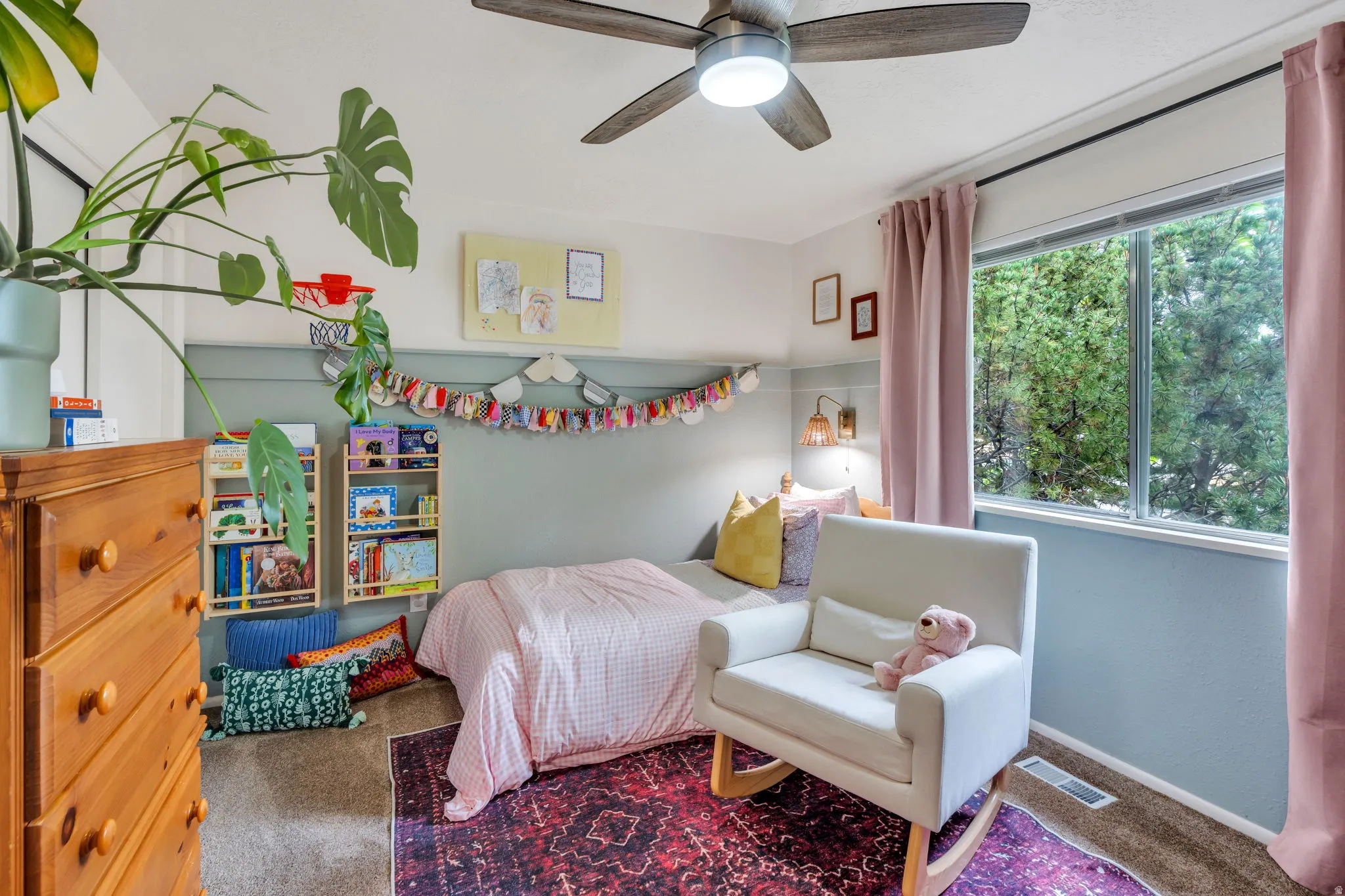Carpeted room featuring a ceiling fan with integrated lighting, a large window with exterior foliage, and a two-tone wall treatment with wainscoting