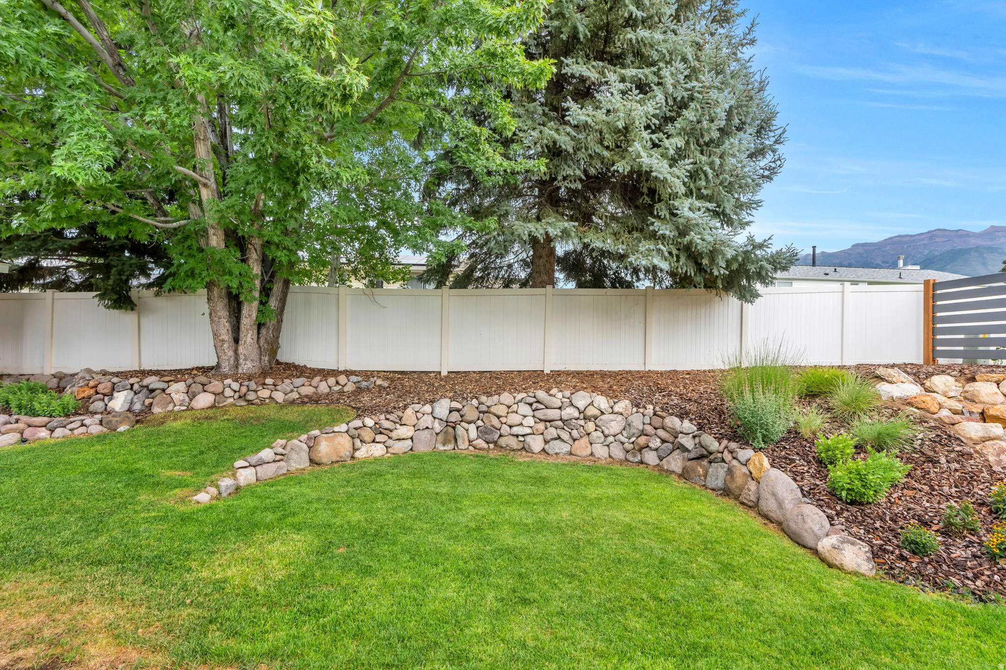 Expansive yard featuring a manicured lawn, established trees, and a low stone retaining wall