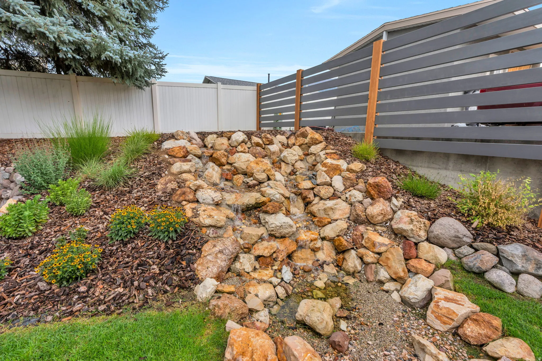 Outdoor rock water feature surrounded by mature landscaping and a privacy fence