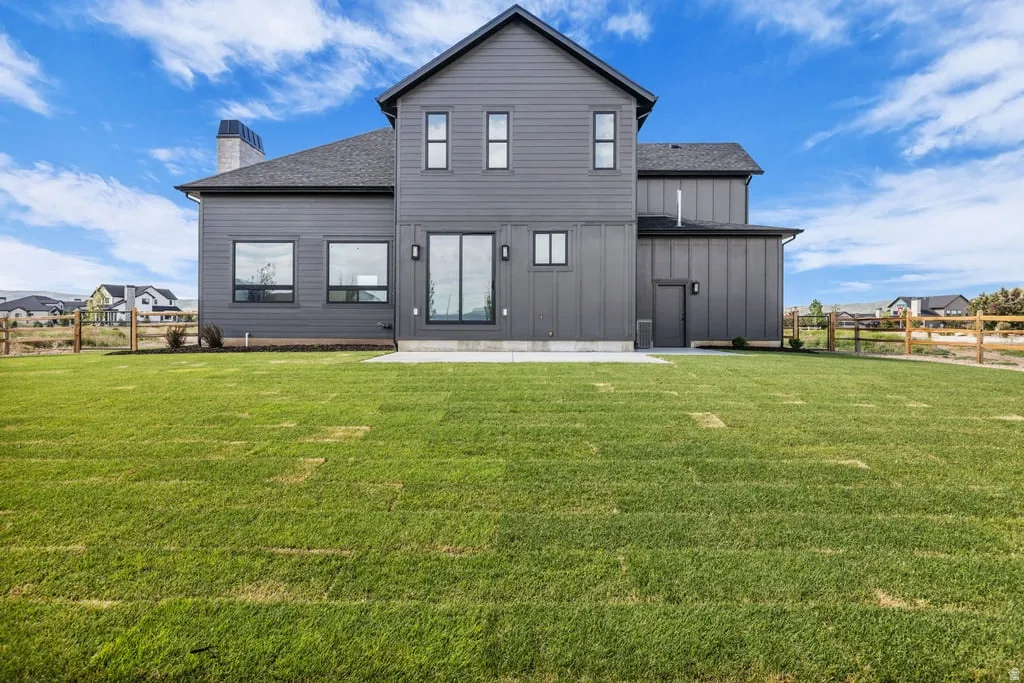 Expansive green lawn with a modern gray exterior featuring board and batten siding, multiple large windows, a sliding glass door, and a concrete patio