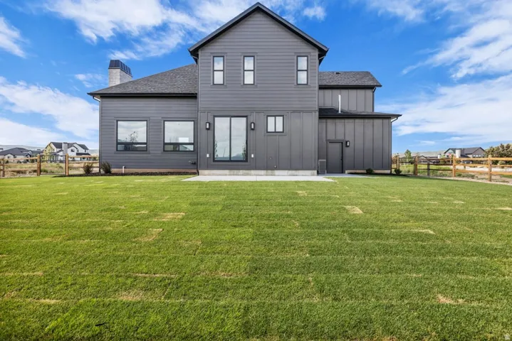 Expansive green lawn with a modern gray exterior featuring board and batten siding, multiple large windows, a sliding glass door, and a concrete patio