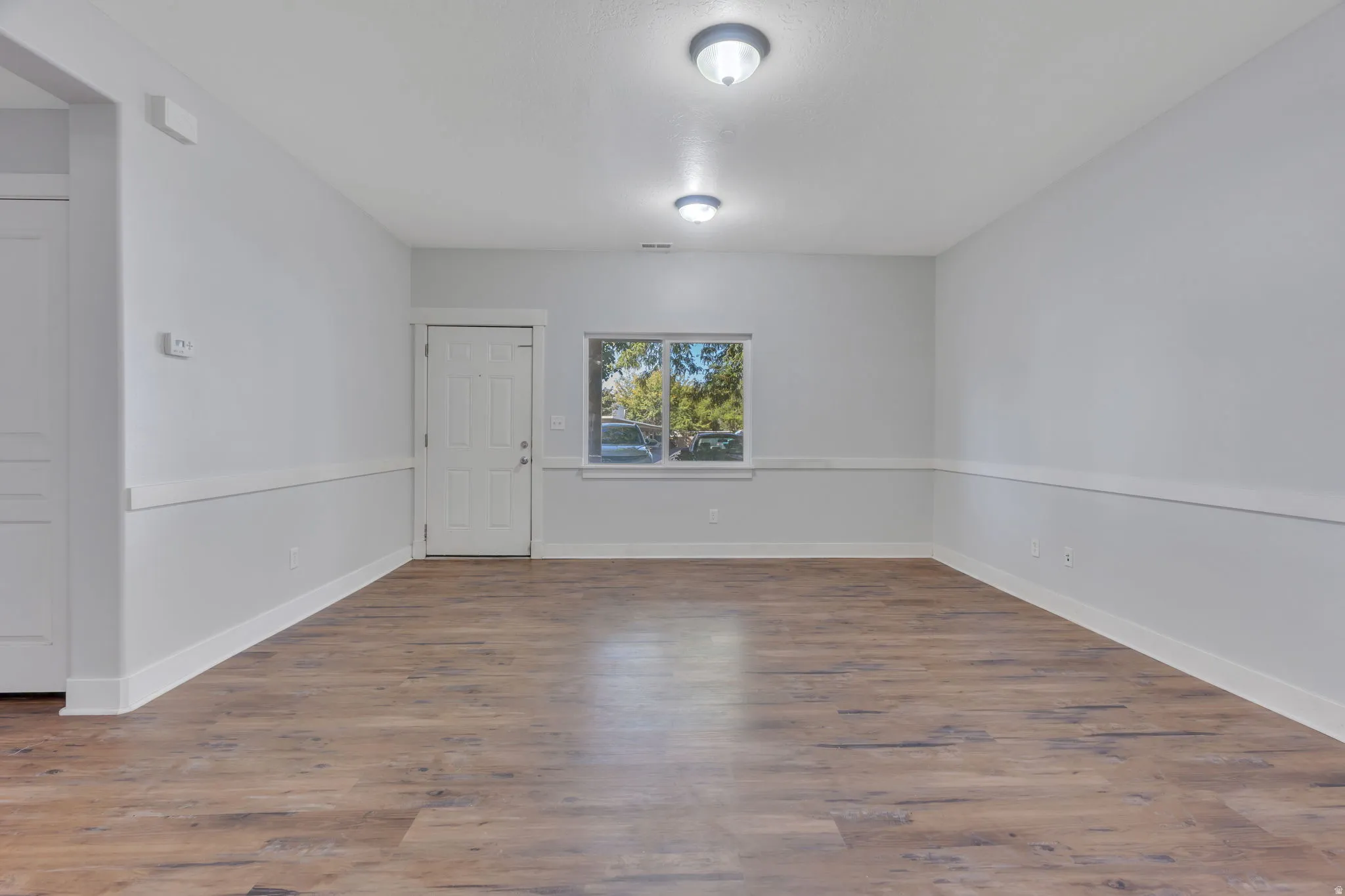 Spacious room featuring wood-finish flooring, neutral paint, a paneled door, and a window with white trim