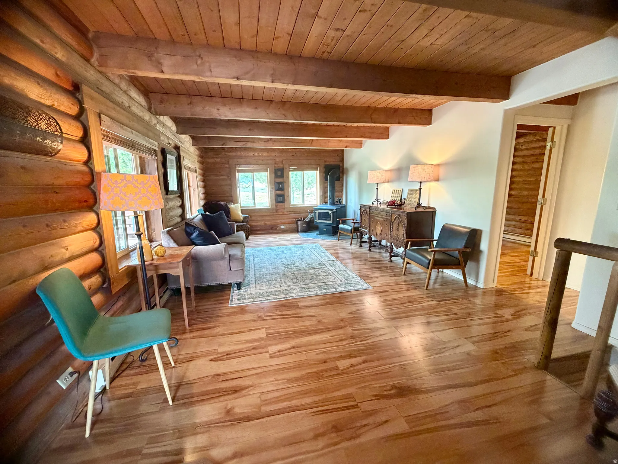 Open-concept living space featuring wood-finish flooring, exposed wood beam ceiling, wood-paneled walls, multiple windows, and a wood-burning stove