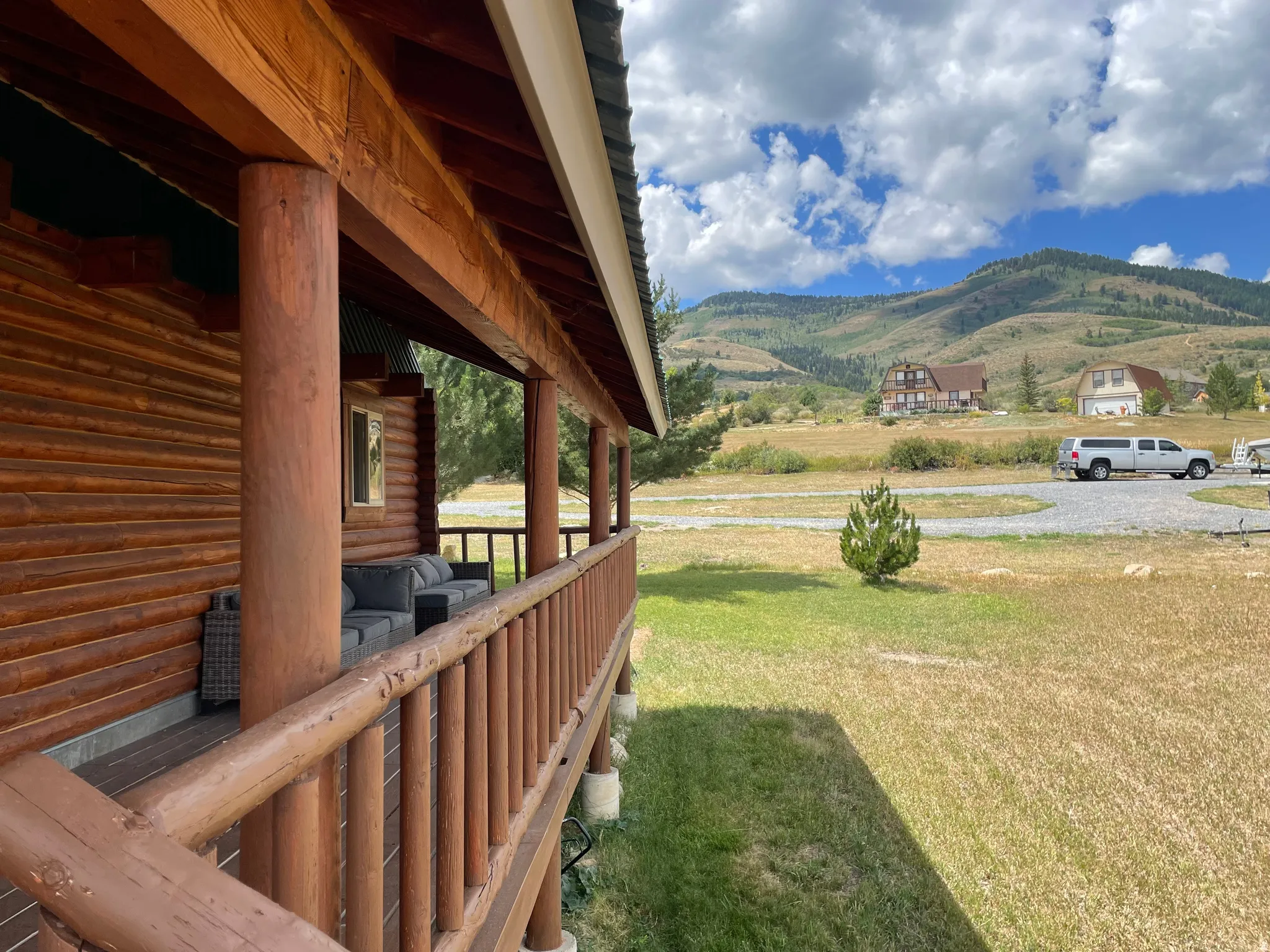 Log cabin exterior with a covered porch featuring wood posts and railing, overlooking a gravel driveway and distant mountainous terrain