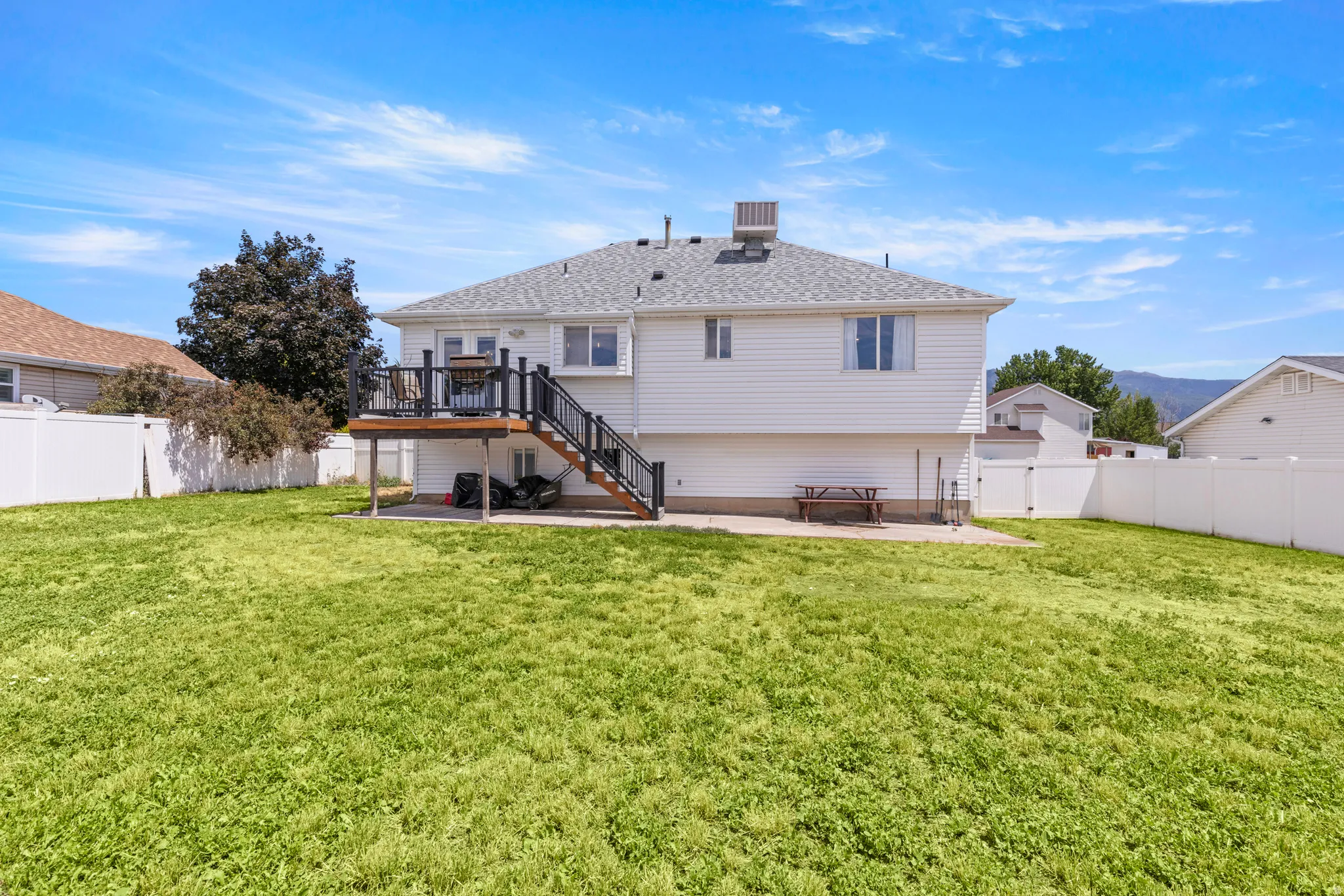 Expansive backyard featuring a large green space, white vinyl fencing, an upper-level wooden deck with black railings, and a lower-level concrete patio