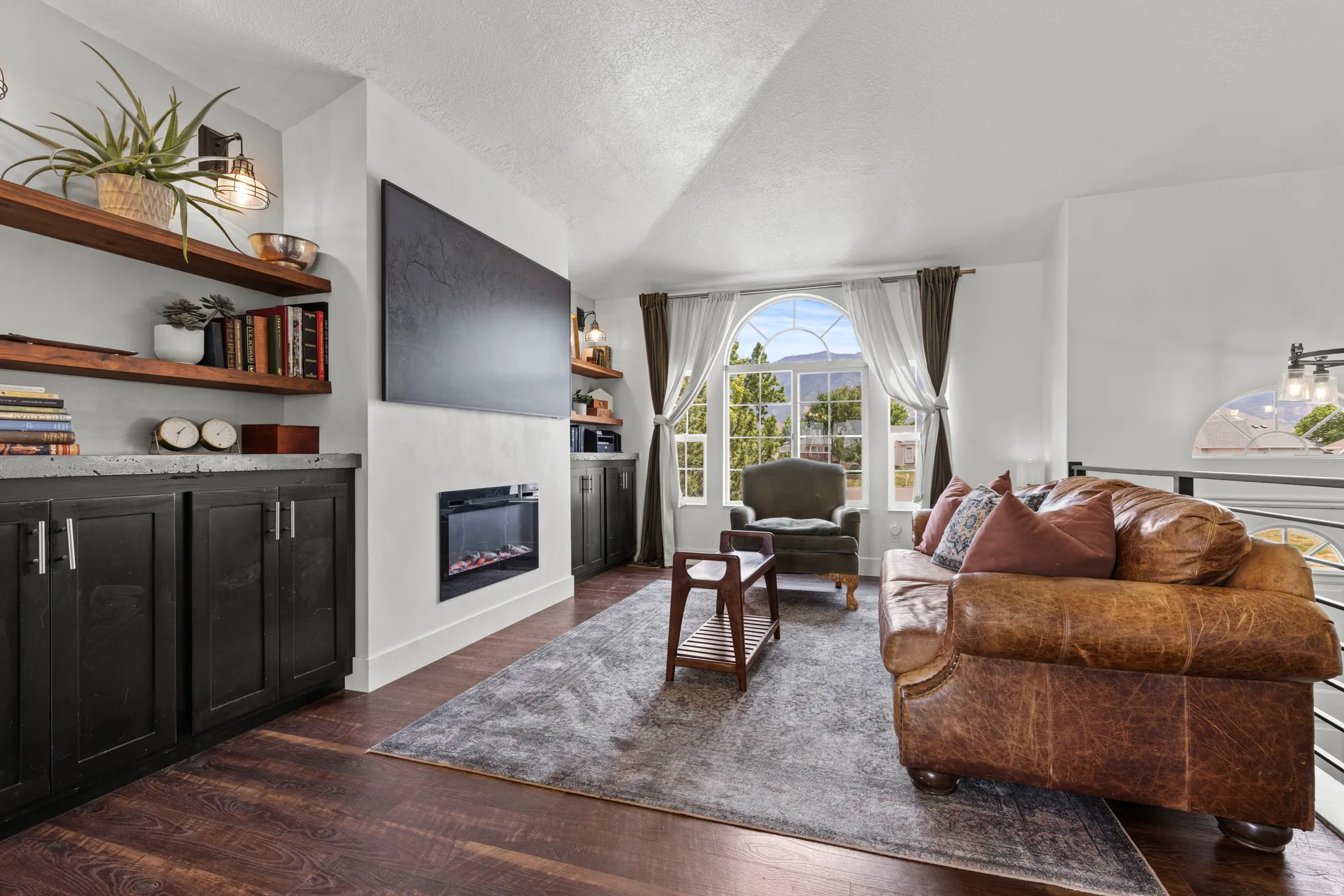 Living area featuring wood-finish flooring, built-in shelving with base cabinetry, an electric fireplace, and a large arched window