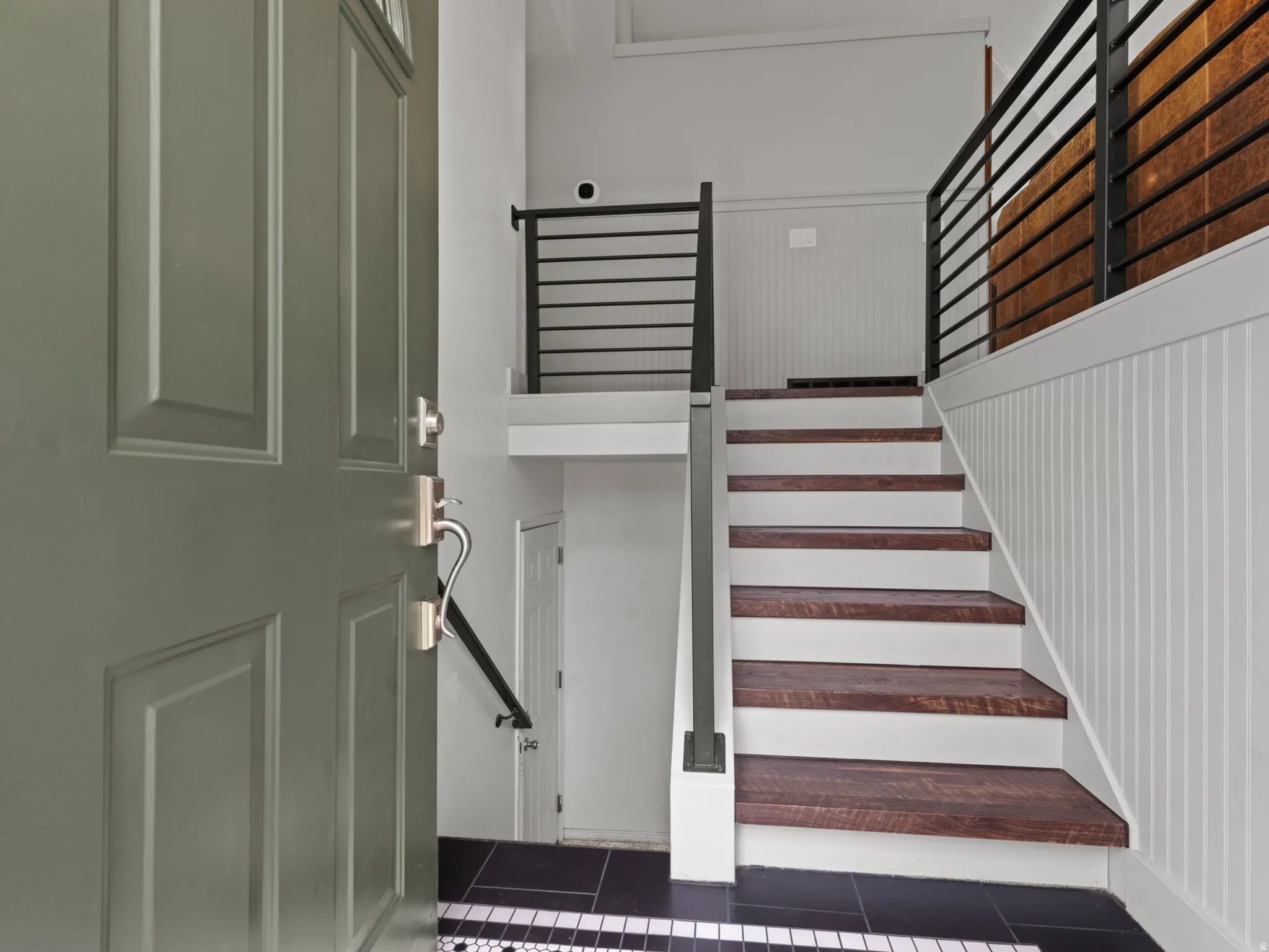 Entryway featuring a paneled door, black and white mosaic tile flooring, wood-finish stair treads, white beadboard paneling, and black metal railings