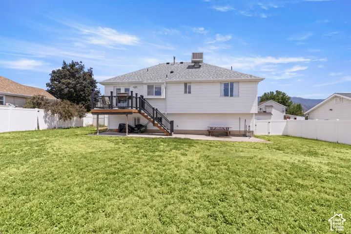 Expansive backyard featuring a large green space, white vinyl fencing, an upper-level wooden deck with black railings, and a lower-level concrete patio