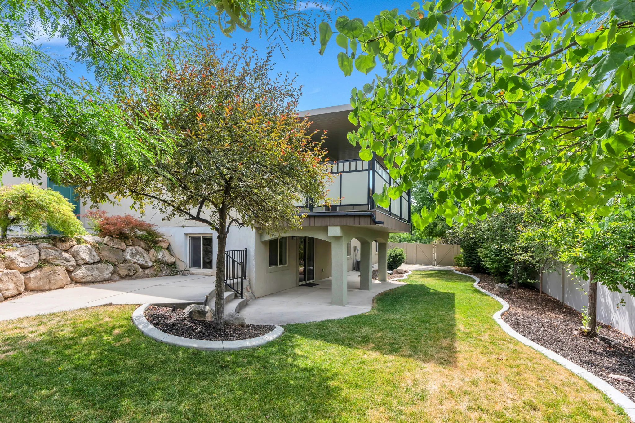 Expansive backyard featuring a manicured lawn, concrete patio, a rock retaining wall, and mature trees