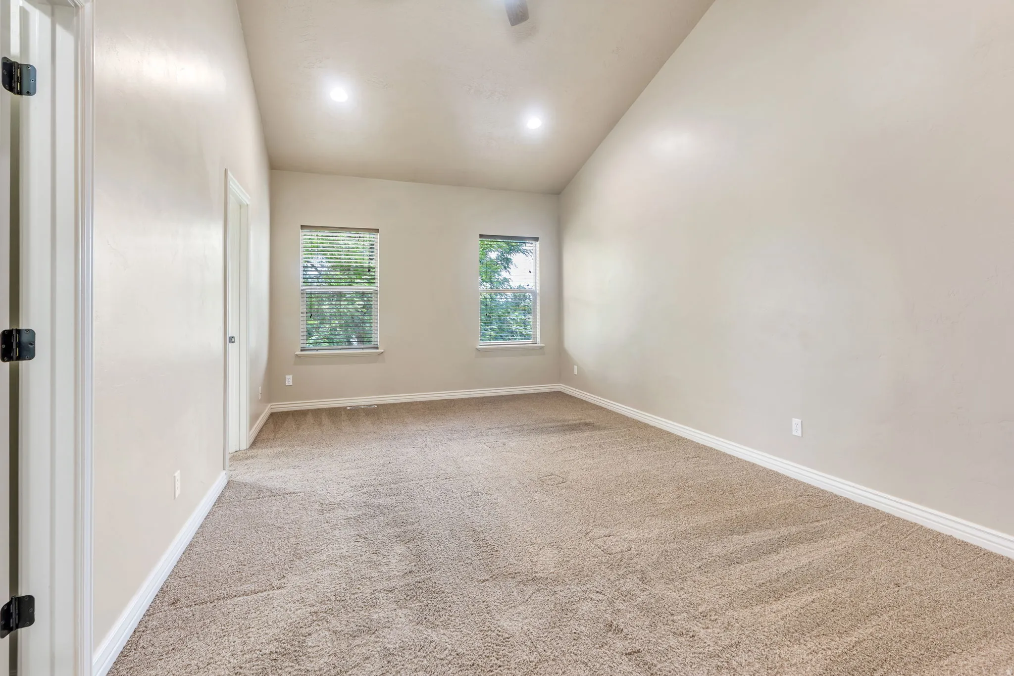 Vaulted ceiling, recessed lighting, two windows with blinds, light-toned carpet, and white trim