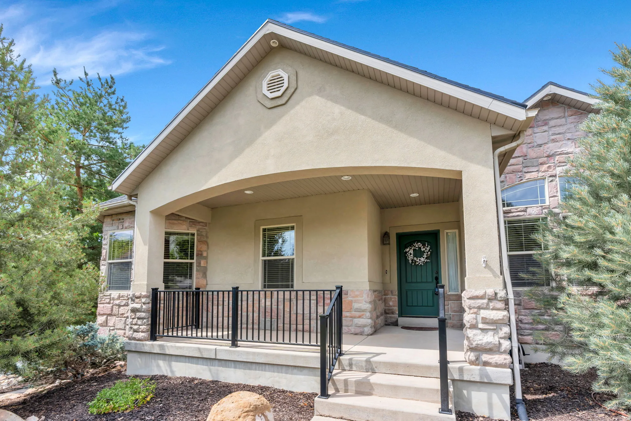 Stucco and stone exterior with a covered entry, featuring a dark green front door, multiple windows, and a black wrought iron railing